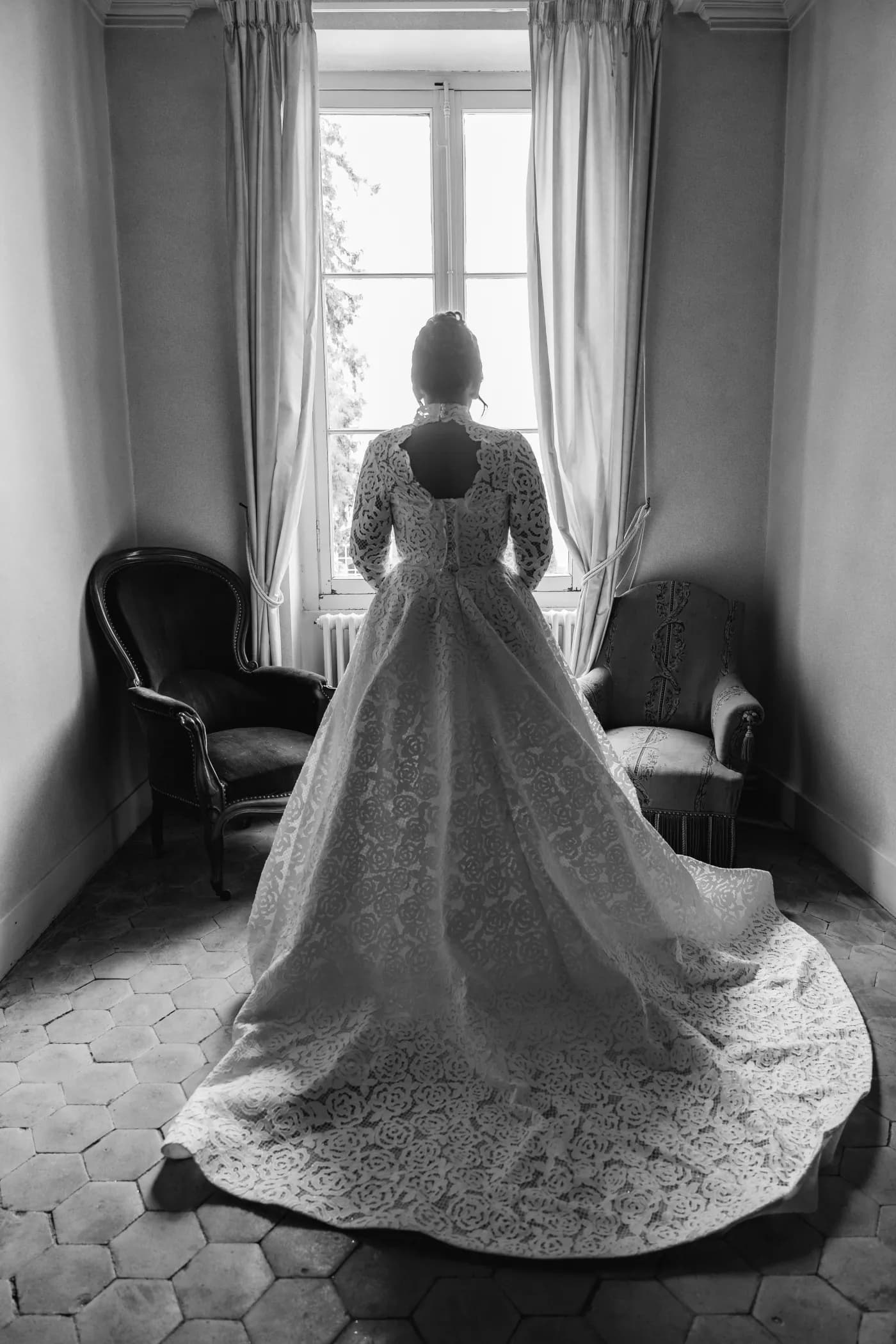 La mariée de dos devant une fenêtre, longue traîne déployée sur le sol, en noir et blanc
